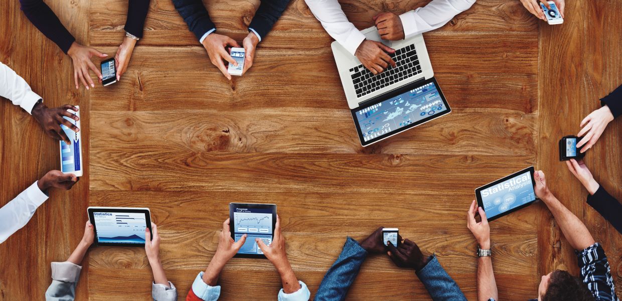 Group of people around a table on different devices, including tablets, laptops, and smartphones